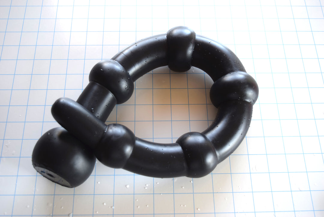 26-inch black silicone Colon Ball enema tube coiled on a blue grid background, showcasing its five large 2.5-inch sensory spheres, thick connecting shaft sections, and 3-inch safety ball flange