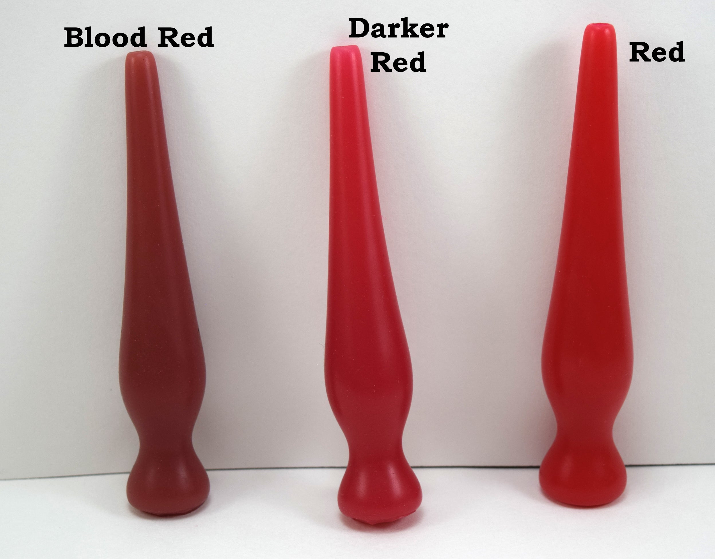 Three long silicone nozzle plugs displayed side-by-side on a white surface to demonstrate color variations. The plugs are labeled from left to right as 'Blood Red', 'Darker Red', and 'Red'; each features an elongated, tapered shape with a rounded base.