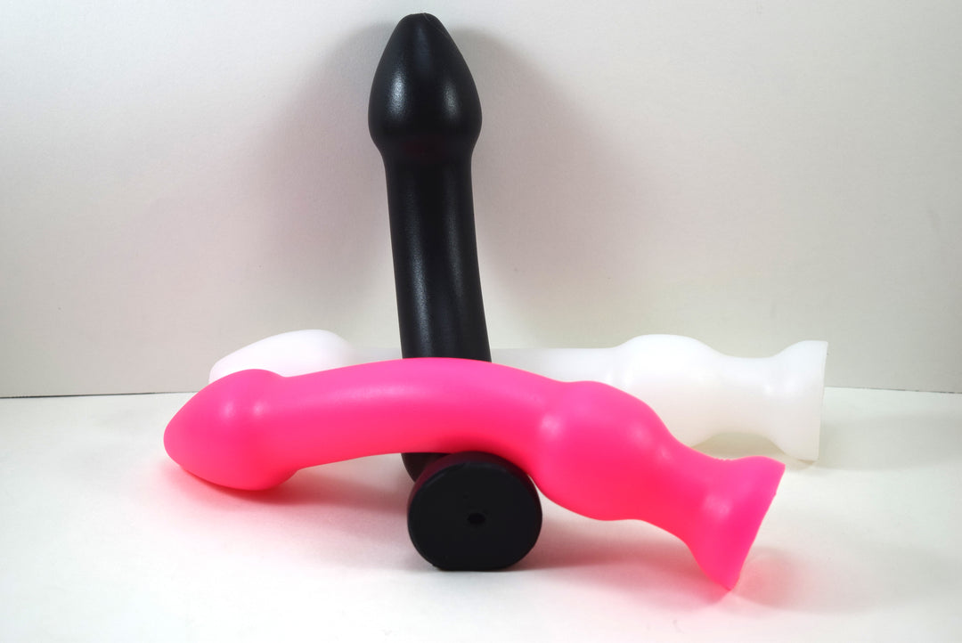 Three DBLBL 1XL silicone colon nozzles shown in solid black, hot pink, and translucent white, showcasing their 2-inch bulbous tips, thick 1.5-inch central shafts, 2-inch barrel retention balls, and flared safety bases