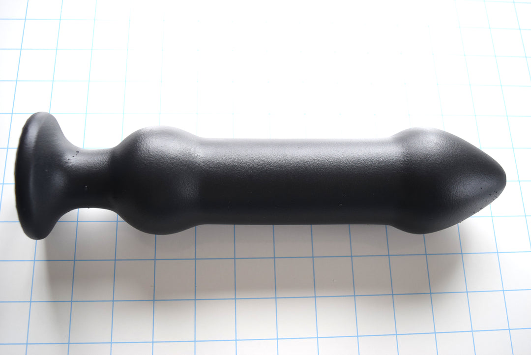 DBLBL 3XL massive black silicone colon nozzle showcasing its colossal 2.5-inch bulbous tip, thick 2.0-inch smooth central shaft, 2.5-inch secondary retention ball, and wide flared safety flange base on a blue grid background