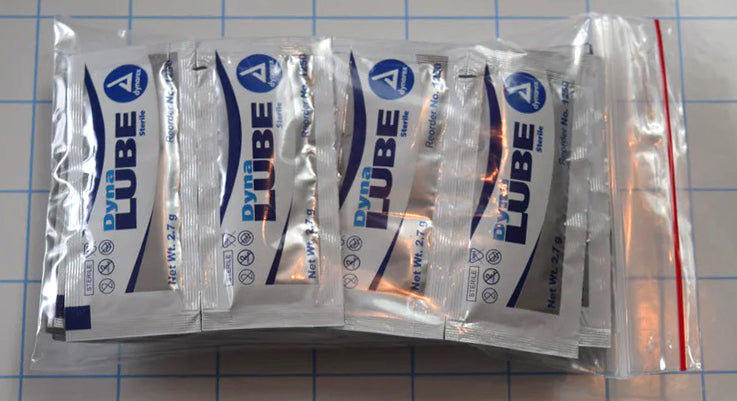 25 2.7g packets of Dynalube sterile lubricating jelly packaged in a clear zip bag on a grid background.
