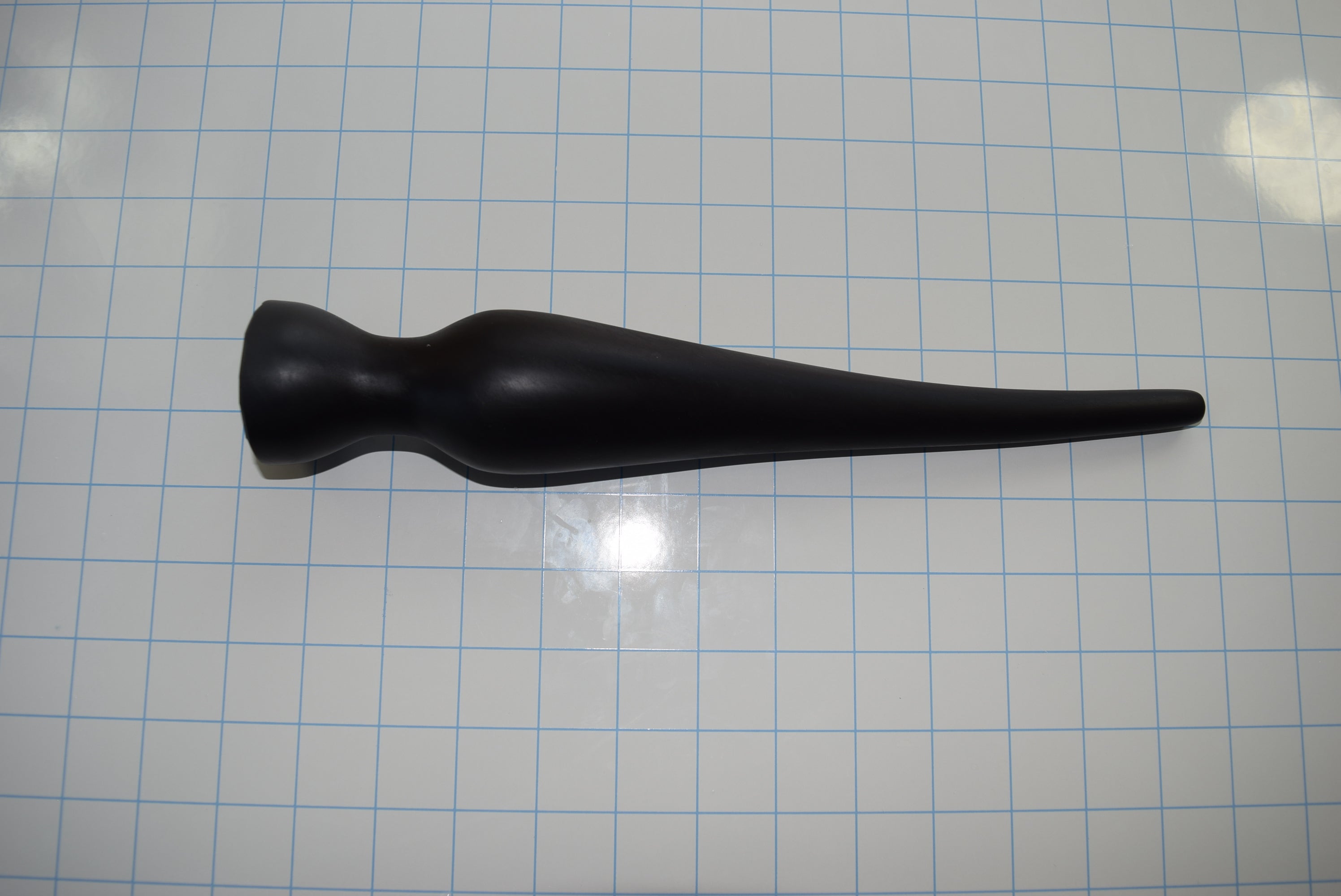 Black EXT CON 2.0 inch silicone enema plug lying horizontally on a blue measurement grid background.
