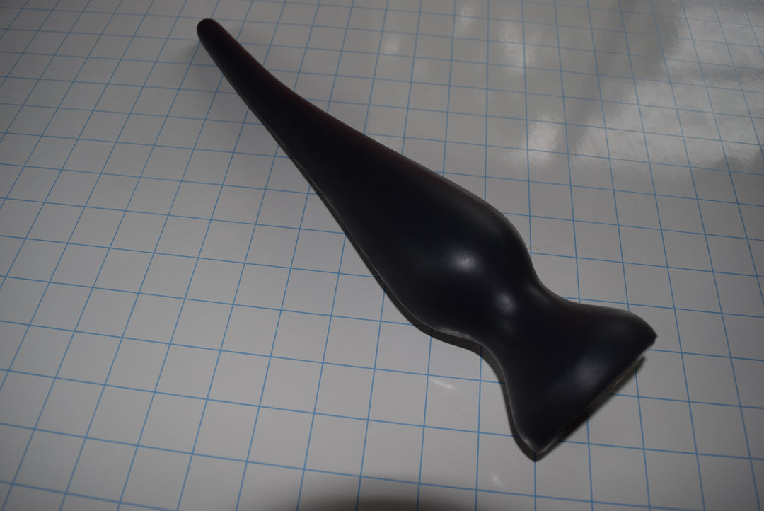 Angled top-down view of a black EXT CON 2.5 inch silicone enema plug resting on a grid surface.