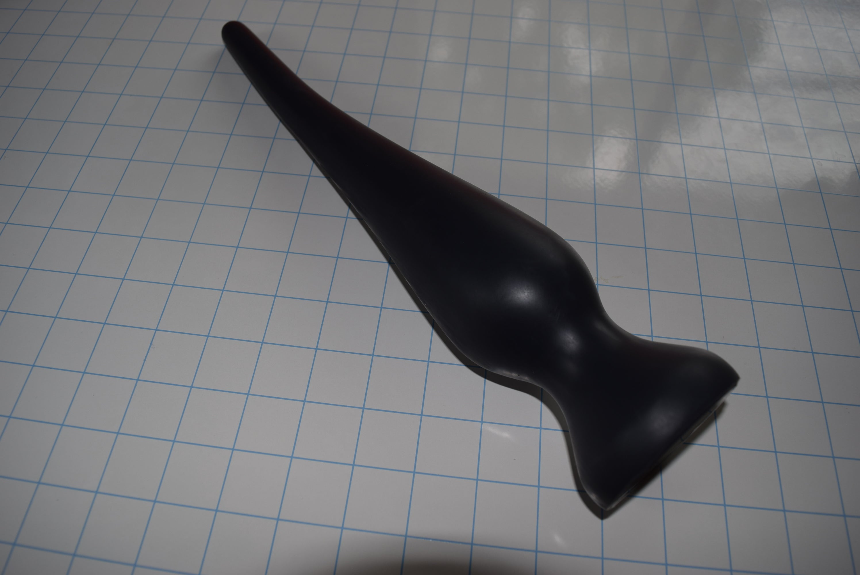 Angled top-down view of a black EXT CON 2.5 inch silicone enema plug resting on a grid surface.