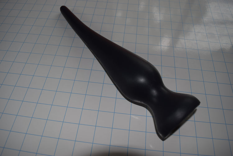 Angled top-down view of a black EXT CON 2.5 inch silicone enema plug resting on a grid surface.