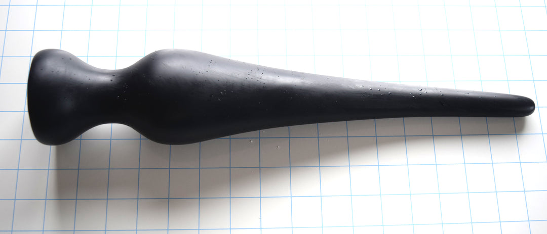 Black EXT CON 3.0 inch silicone enema retention plug lying horizontally on a blue measurement grid.