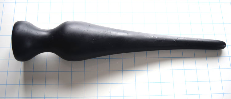 Black EXT CON 3.0 inch silicone enema retention plug lying horizontally on a blue measurement grid.