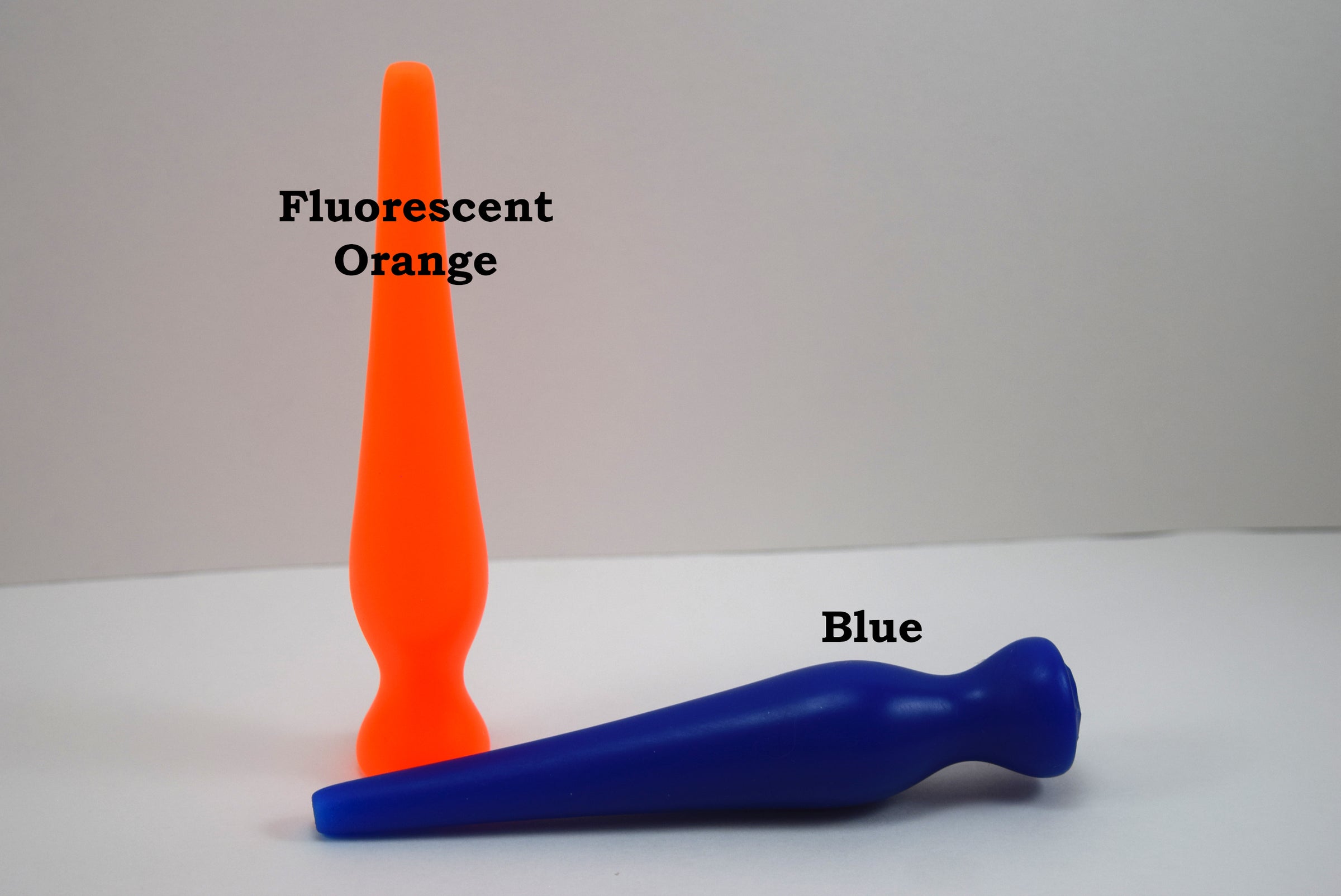 Two long silicone nozzle plugs shown against a white background to illustrate color differences. The plug standing vertically is labeled 'Fluorescent Orange', and the plug lying horizontally is labeled 'Blue'; both feature an elongated, tapered shape.