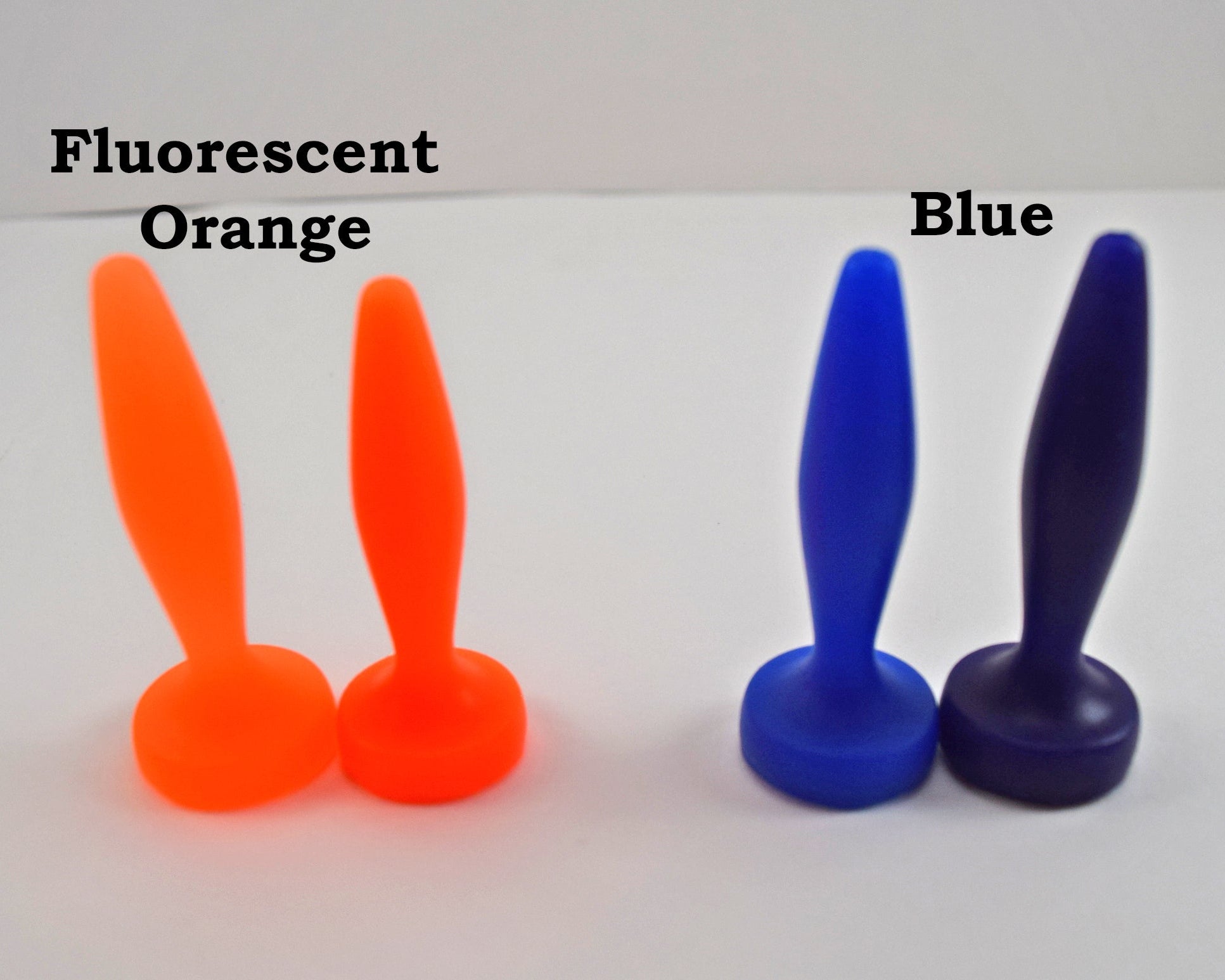 Two pairs of silicone urethral sounds displayed on a white surface to show color differences. The pair on the left is labeled 'Fluorescent Orange', and the pair on the right is labeled 'Blue'; all feature a diamond-shaped body, tapered tip, and flat base