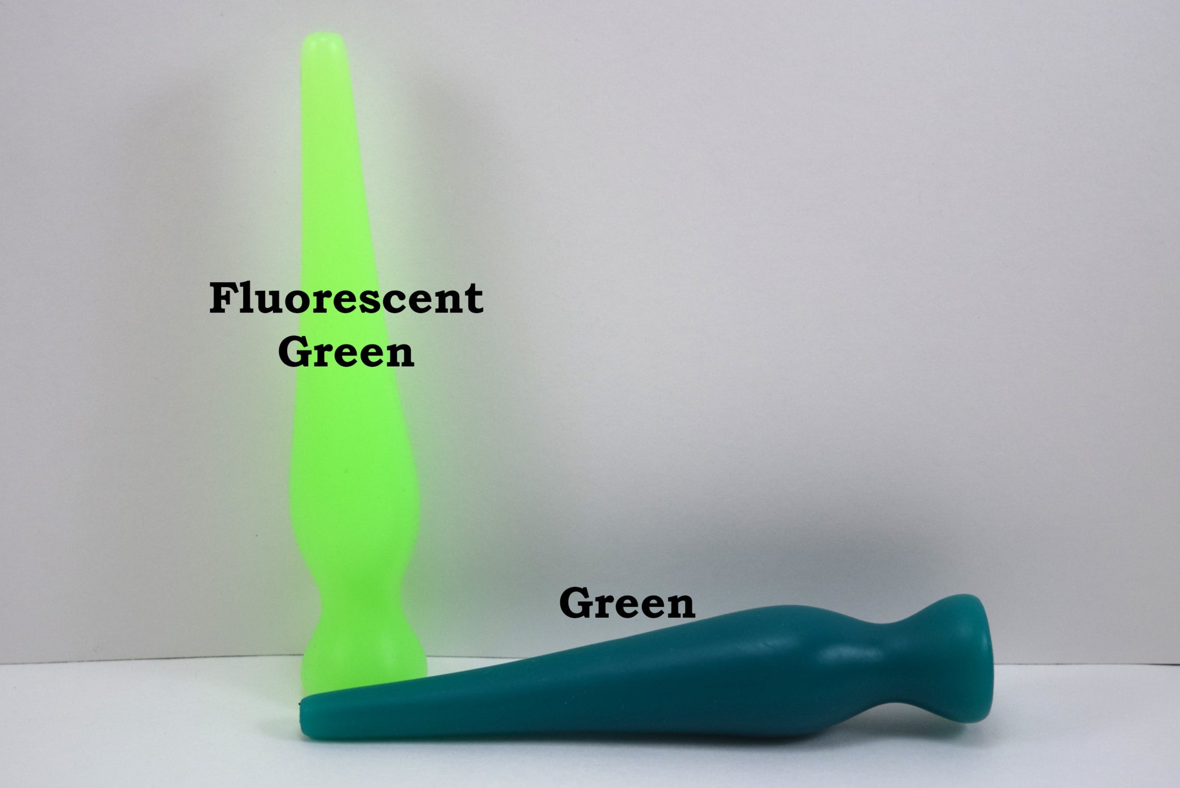 Two long silicone nozzle plugs shown against a white background to illustrate color differences. The plug standing vertically is labeled 'Fluorescent Green', and the plug lying horizontally is labeled 'Green'; both feature an elongated, tapered shape