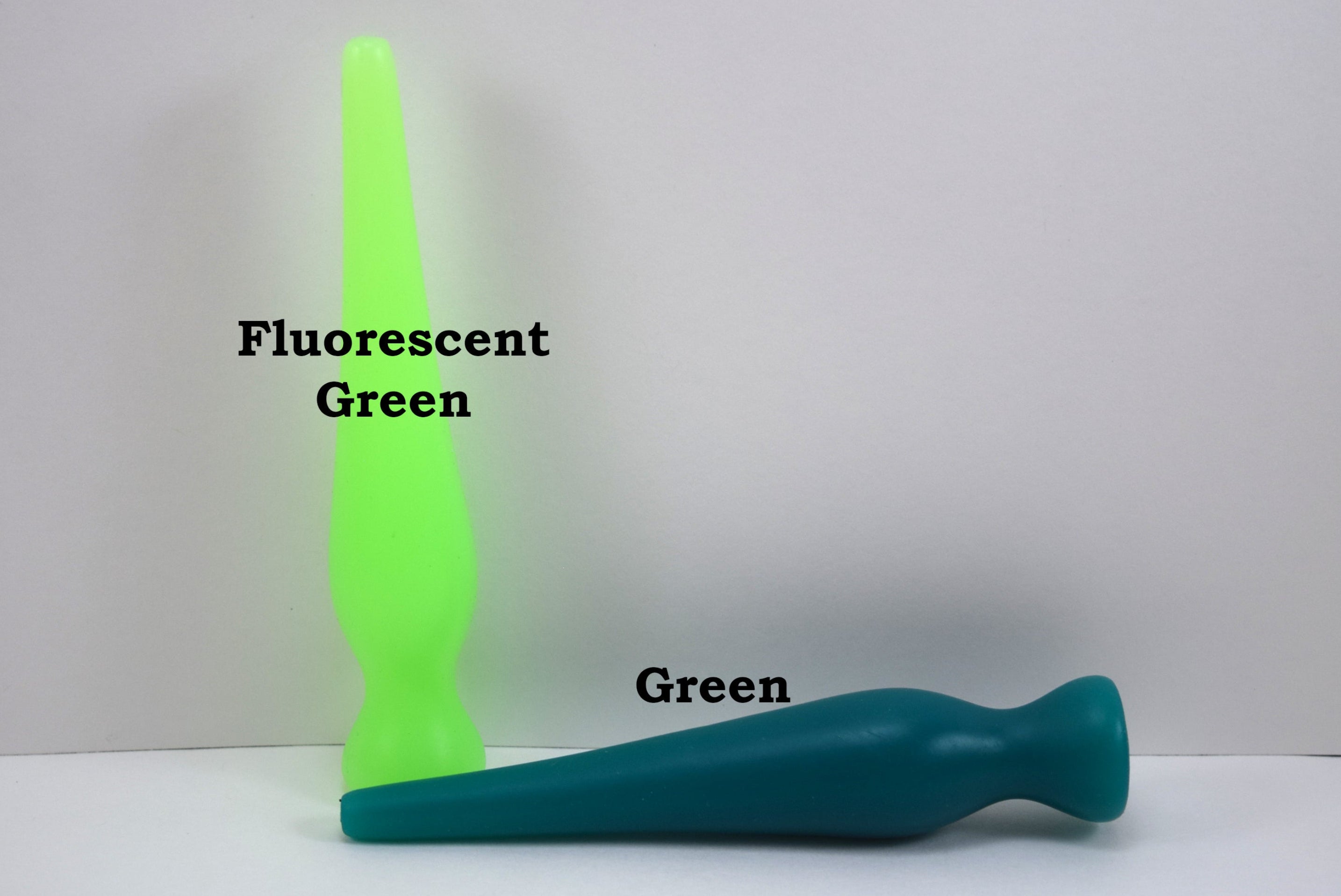 Two long silicone nozzle plugs shown against a white background to illustrate color differences. The plug standing vertically is labeled 'Fluorescent Green', and the plug lying horizontally is labeled 'Green'; both feature an elongated, tapered shape