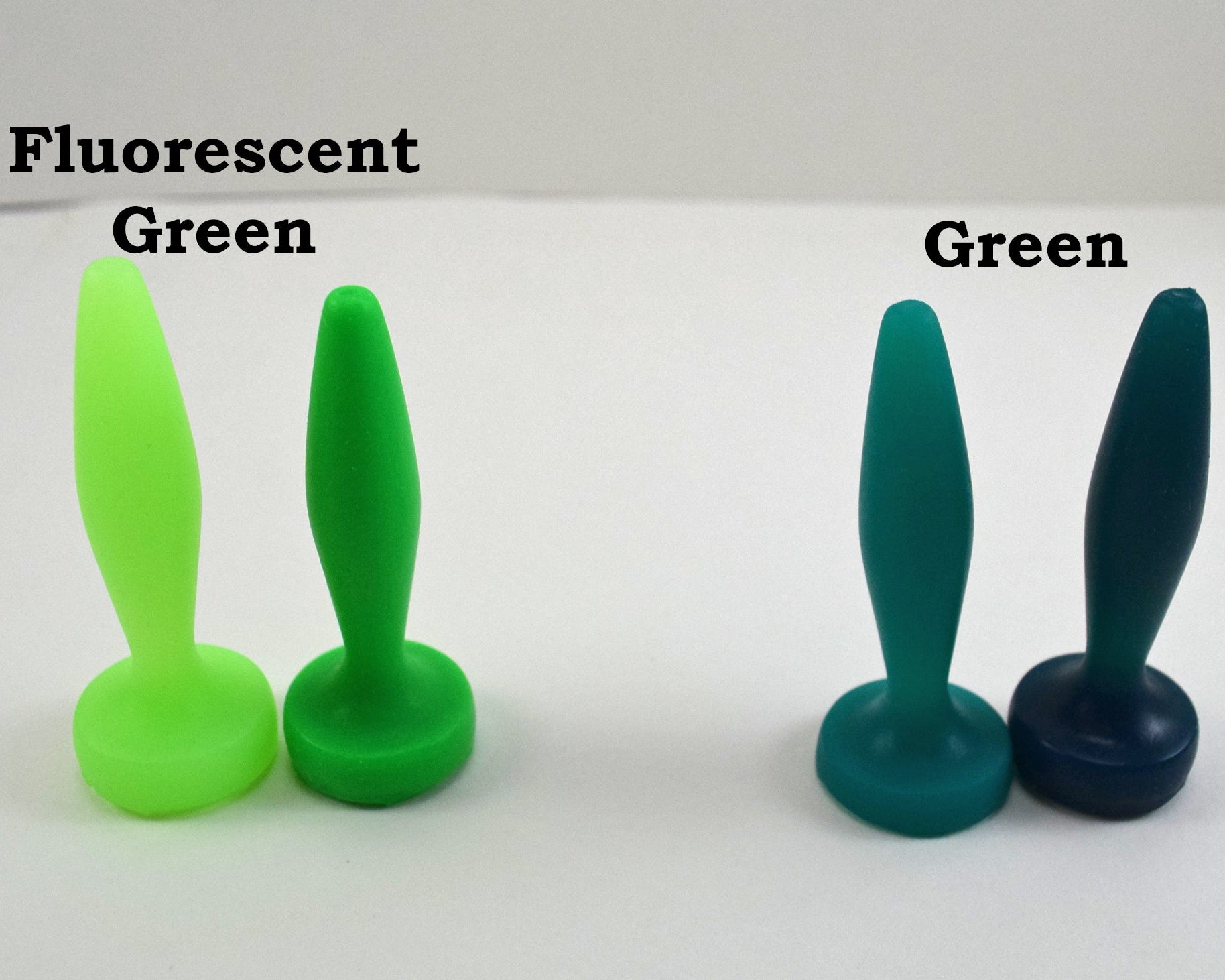 Two pairs of silicone urethral sounds displayed on a white surface to show color differences. The pair on the left is labeled 'Fluorescent Green', and the pair on the right is labeled 'Green'; all feature a diamond-shaped body, tapered tip, and flat base