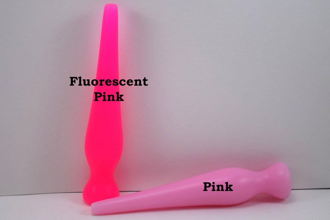 Two long silicone nozzle plugs shown against a white background to illustrate color differences. The plug standing vertically is labeled 'Fluorescent Pink', and the plug lying horizontally is labeled 'Pink'; both feature an elongated, tapered shape.