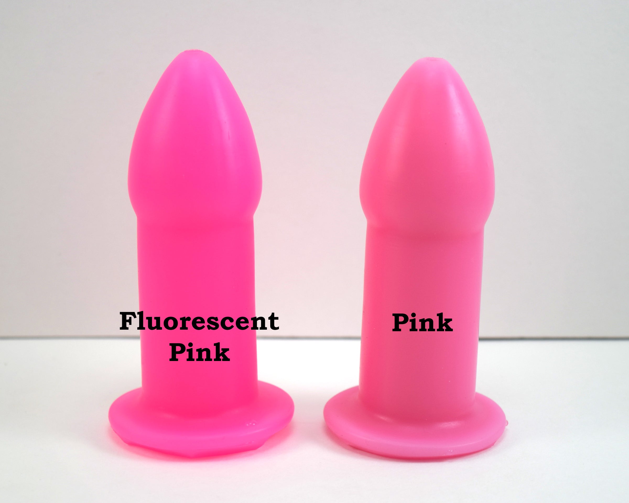 Two silicone nozzle plugs side-by-side on a white surface, demonstrating color differences. The plug on the left is labeled 'Fluorescent Pink', and the plug on the right is labeled 'Pink'; both have a tapered tip and wide safety flange
