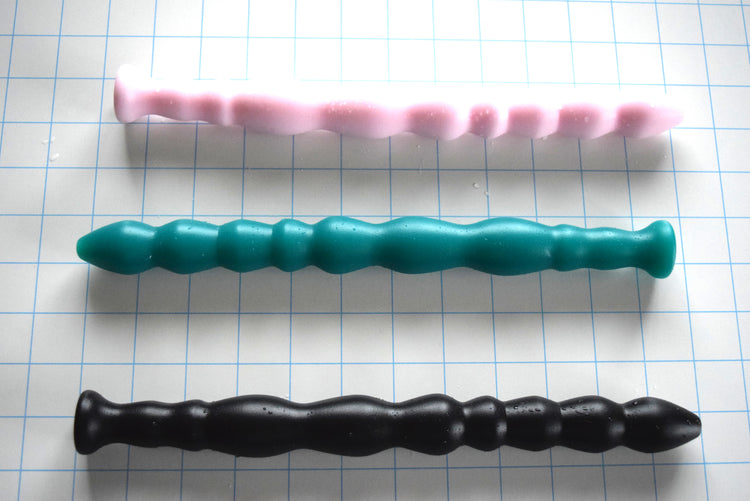 Three Knight textured silicone enema nozzles in pink, teal-green, and black on a blue grid background, showcasing the 9-bead undulating shaft profile, 1.1-inch maximum diameter spheres, and flared safety bases