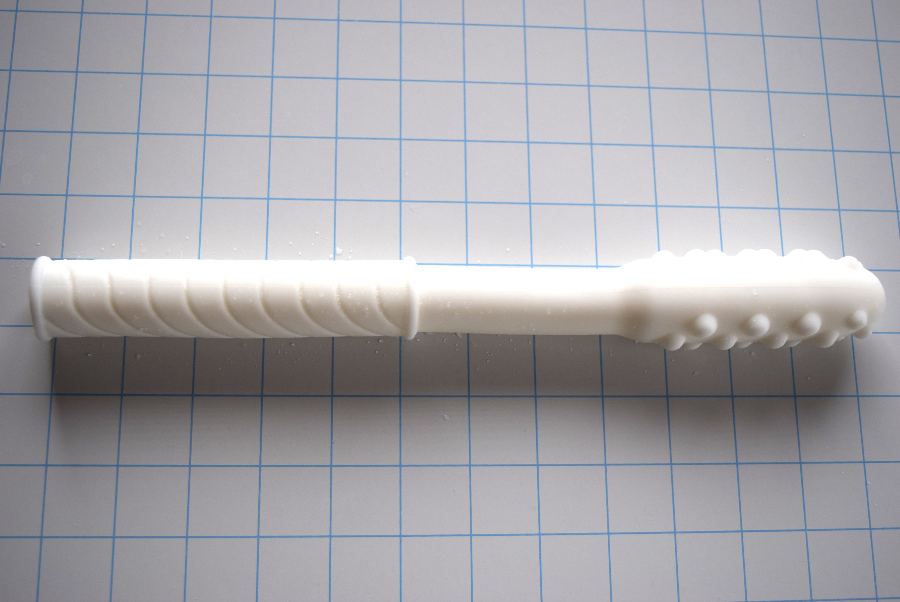 White Knobby Tip silicone enema nozzle with textured bumps and diagonal grip shaft lying horizontally on a blue measurement grid.