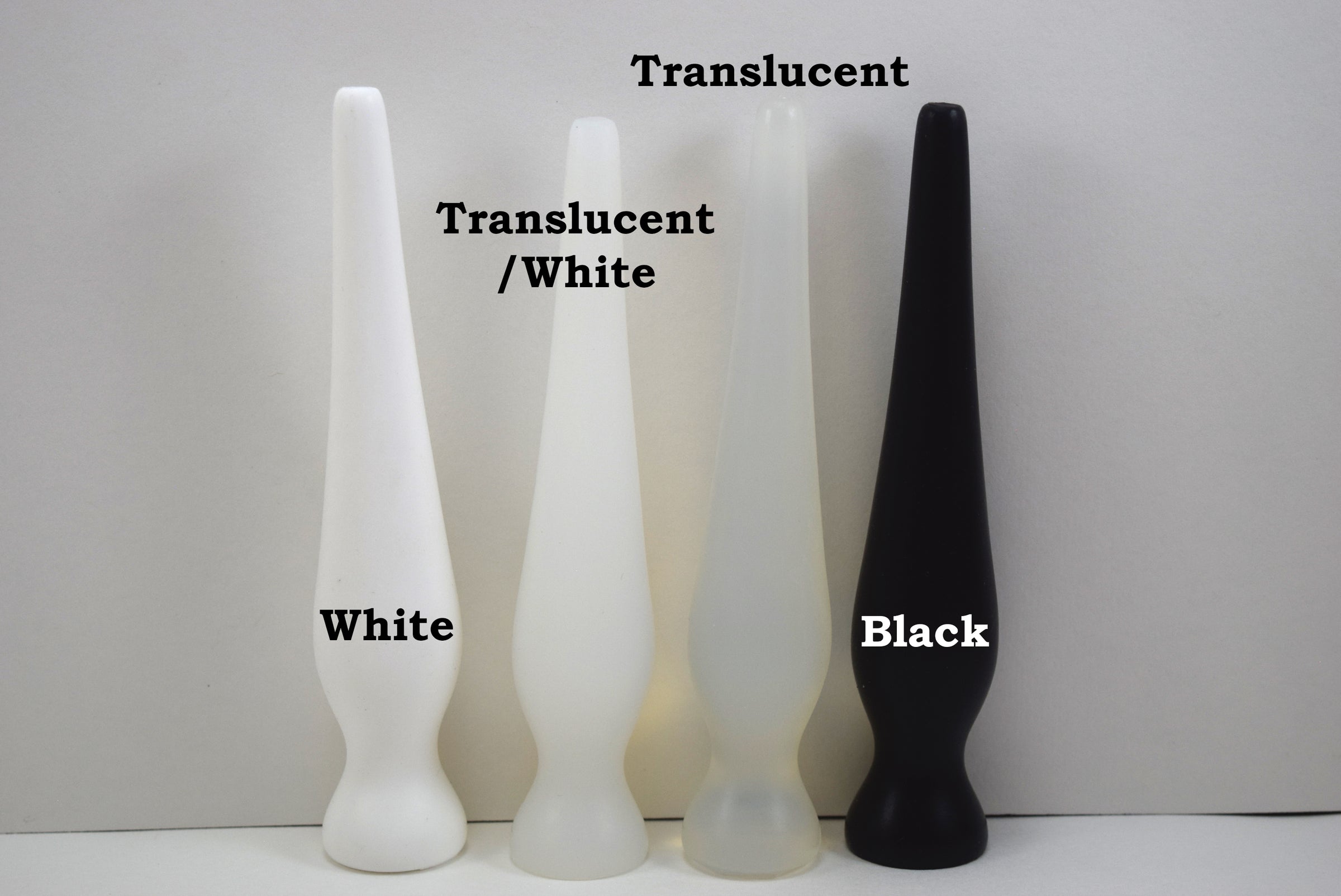 Four long silicone nozzle plugs displayed in a row on a white background to show color variations. From left to right, the plugs are labeled 'White', 'Translucent/White', 'Translucent', and 'Black'; all feature an elongated, tapered shape with a rounded base.