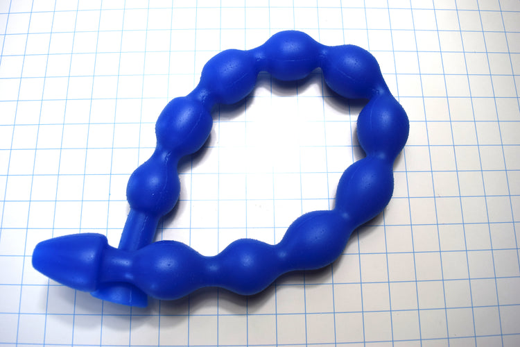 MAXIBALL 30-inch blue silicone colon tube showing ten segmented 1.8-inch retention bulbs, a tapered tip, and a 1.9-inch safety flange coiled on a blue grid background.