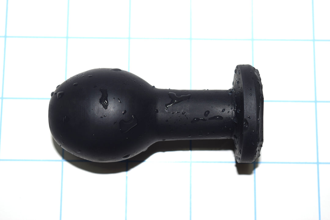 Model E compact black silicone enema nozzle with water droplets on its surface, showcasing its 1.4-inch ball tip, short 3/4-inch narrow shaft, and 2.8-inch total insertable length on a blue grid background.