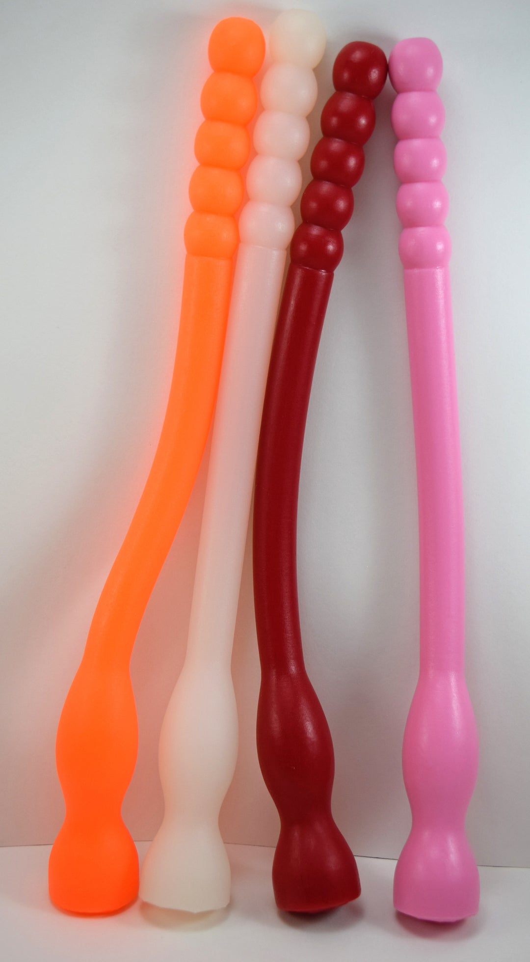 Four Model F flexible silicone colon tubes shown in orange, translucent-white, red, and pink, showcasing the 5-bead rolling ribbed tips, long smooth shafts, and integrated base retaining balls