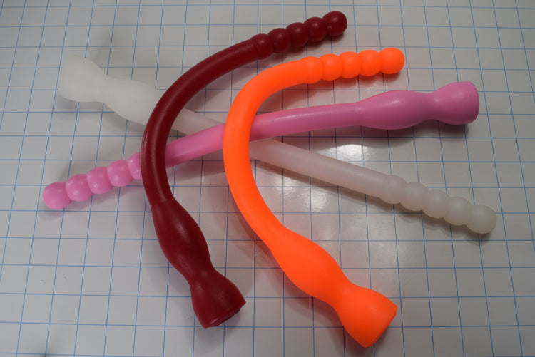 Group of flexible Model F silicone colon nozzles in pink, white, dark red, and orange bent into curves across a grid background.