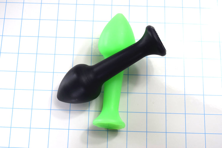 Black and fluorescent green Model Y silicone enema nozzles crossed over each other on a blue measurement grid.