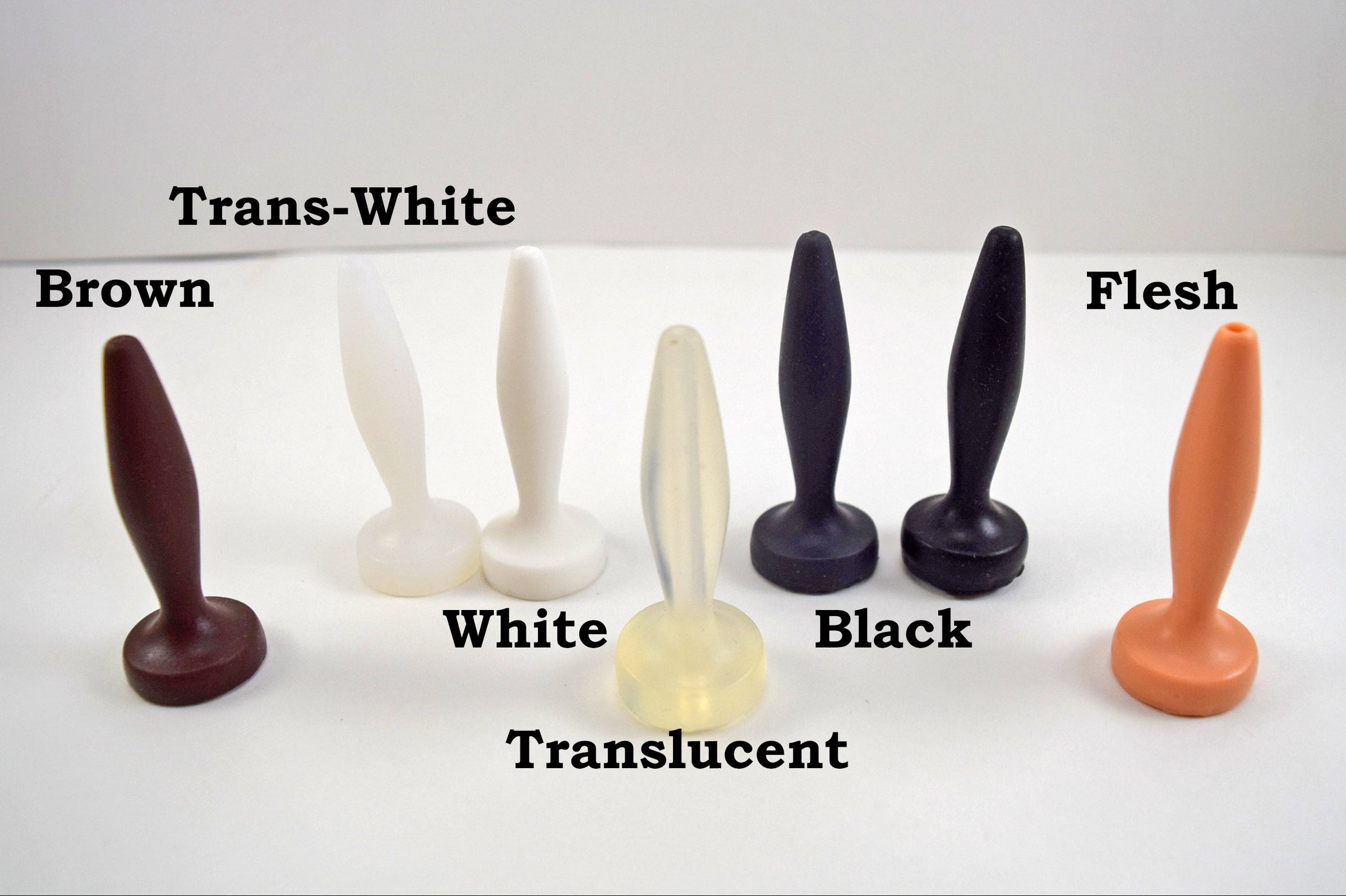 A collection of silicone nozzle plugs displayed on a white surface, showcasing a variety of available colors. The plugs are labeled from left to right as 'Brown', 'Trans-White', 'White', 'Translucent', 'Black', and 'Flesh', each featuring a tapered tip and wide base