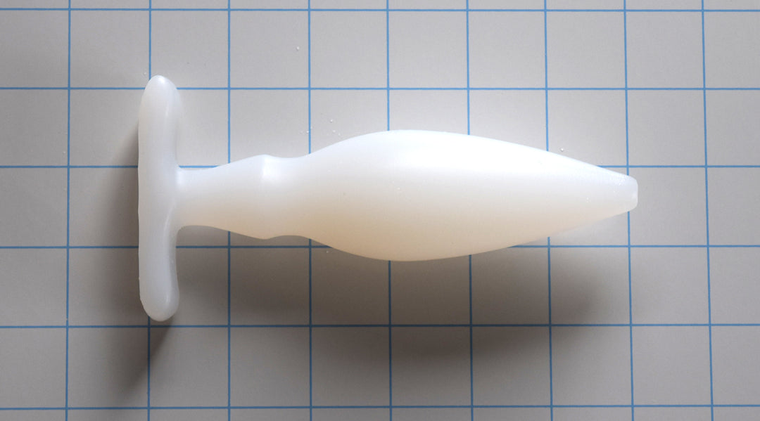 OF 1.6 translucent white silicone enema nozzle on a blue grid background, showcasing its 0.5-inch rounded tip, gradual 1.6-inch taper, dual-stage retention valleys, and a 3-inch wide contoured oval base flange