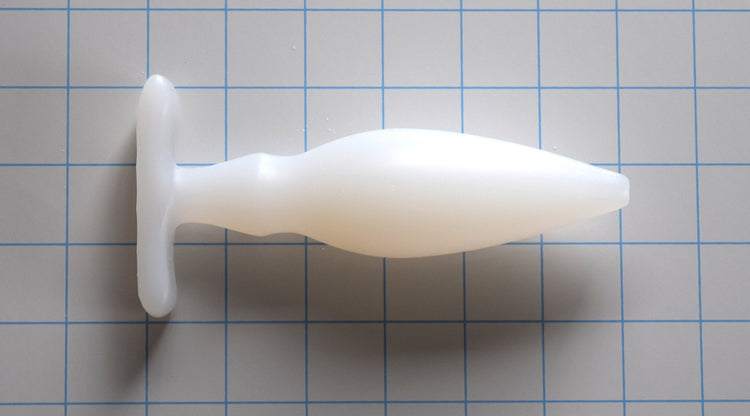 OF 1.6 translucent white silicone enema nozzle on a blue grid background, showcasing its 0.5-inch rounded tip, gradual 1.6-inch taper, dual-stage retention valleys, and a 3-inch wide contoured oval base flange