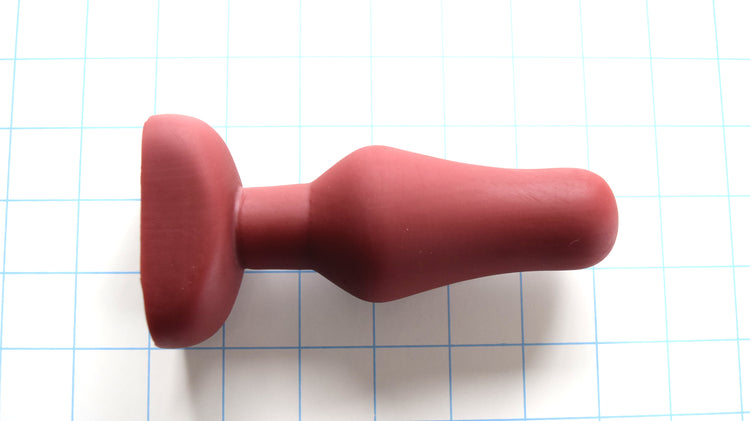 OF 2 medium 6-inch oval flange-style silicone enema nozzle cast in brick red on a white grid background, highlighting the 1.25-inch tip, 1.9-inch maximum swell, and contoured gluteal base flange