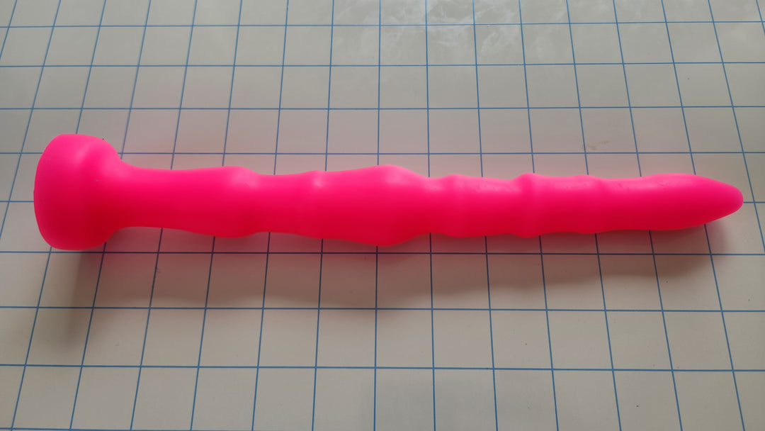 Off-centered 11-inch hot pink silicone enema nozzle and short colon tube displaying its undulating rib pattern, 1.5-inch maximum center rib diameter, and 2-inch safety base flange on a blue grid background