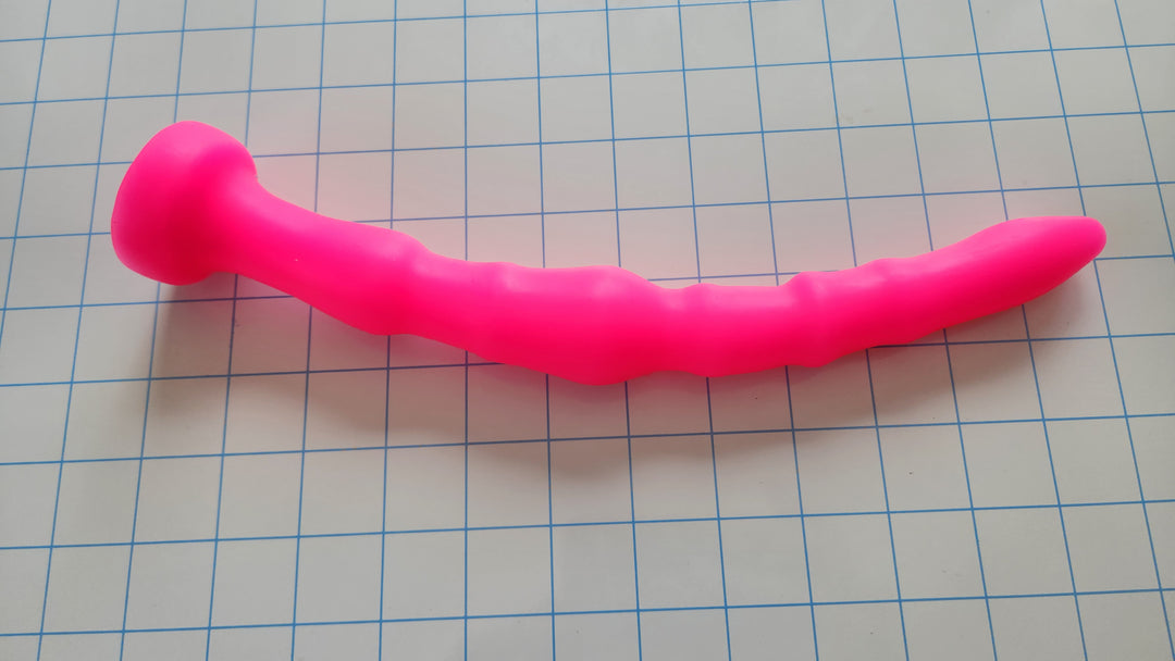 Bright pink Off-Centered 11-inch silicone enema nozzle with asymmetrical undulating ribs lying on a grid paper surface.