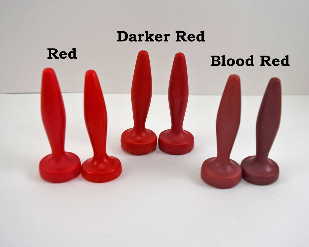 Three pairs of silicone urethral sounds displayed on a white surface to demonstrate color variations. The pairs are labeled from left to right as 'Red', 'Darker Red', and 'Blood Red'; all feature a diamond-shaped body, tapered tip, and flat base.