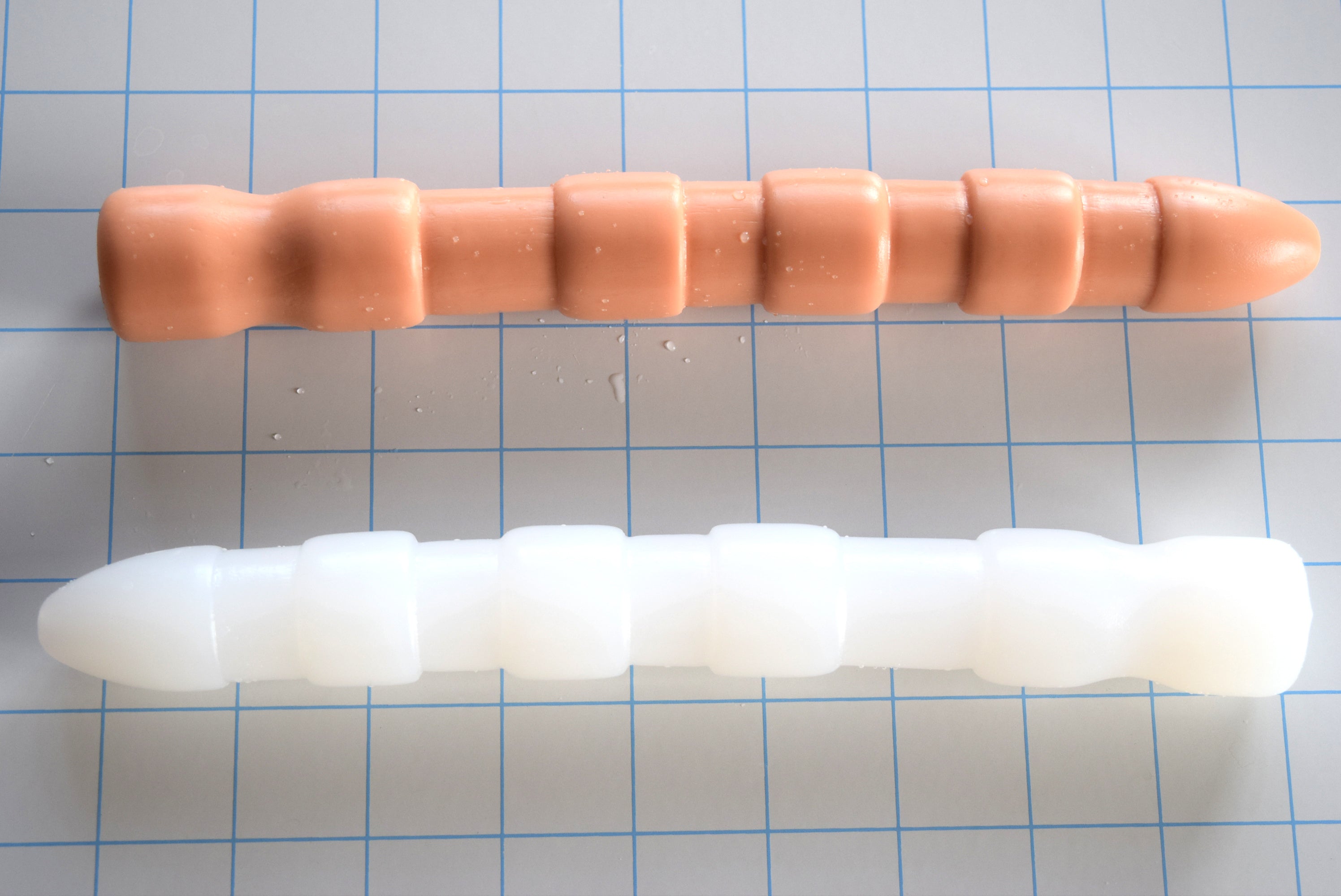 Flesh-tone and translucent white Ribbed Shooter 9-inch silicone enema nozzles displaying 26mm bulbous tip, 28mm sensory ribs, 24mm sphincter valley, and retention flange on a blue grid background.