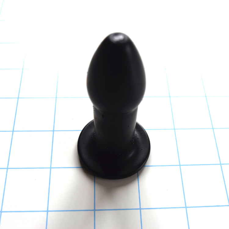 Black silicone rectal training dilator standing upright on a blue grid background.