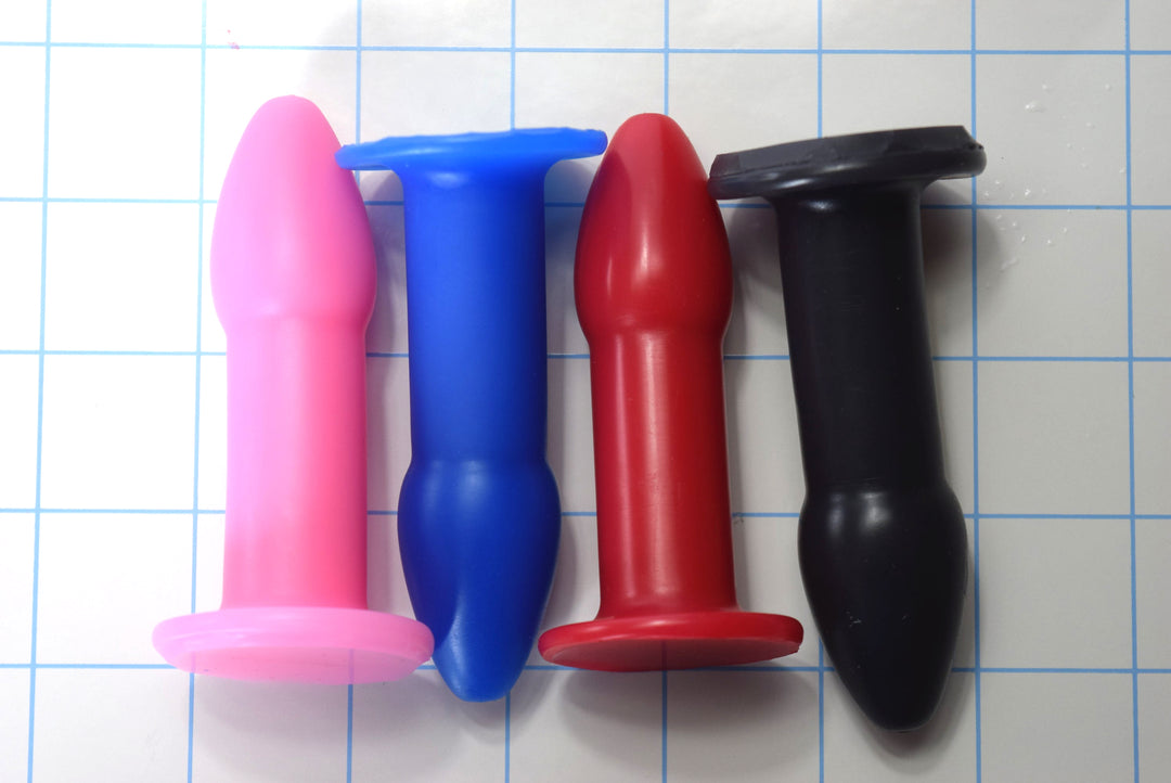 Pink, blue, red, and black 1-inch silicone rectal training dilators arranged side-by-side on a blue grid background.