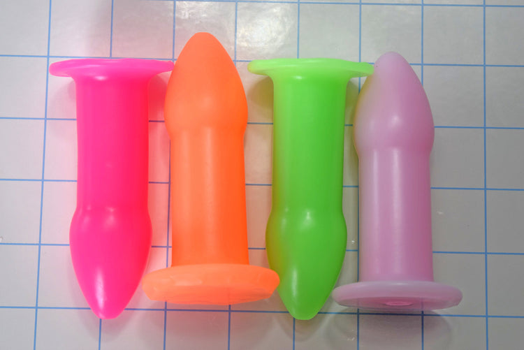 Pink, orange, green, and lavender silicone rectal training dilators arranged on a blue grid background.