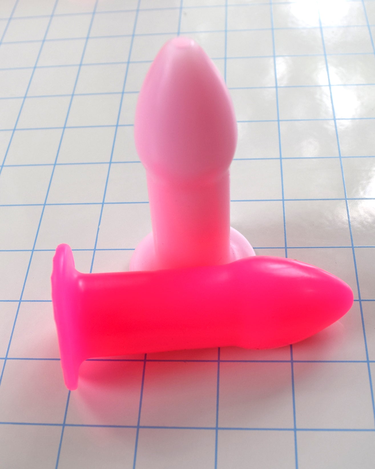 Fluorescent pink and standard pink silicone rectal training dilators placed together on a blue grid background.