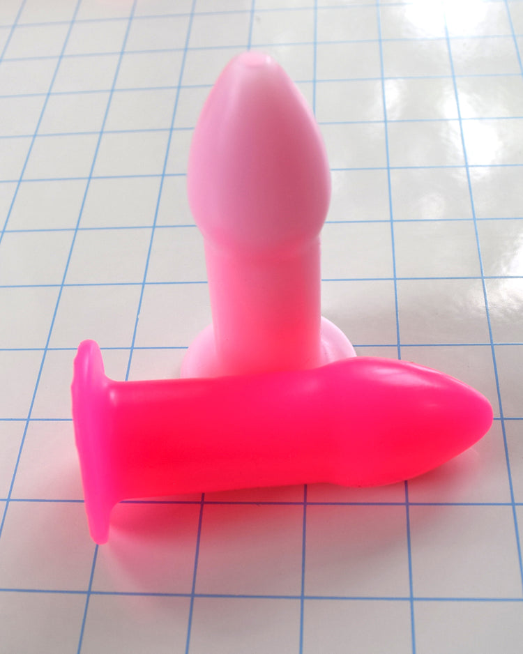 Fluorescent pink and standard pink silicone rectal training dilators placed together on a blue grid background.