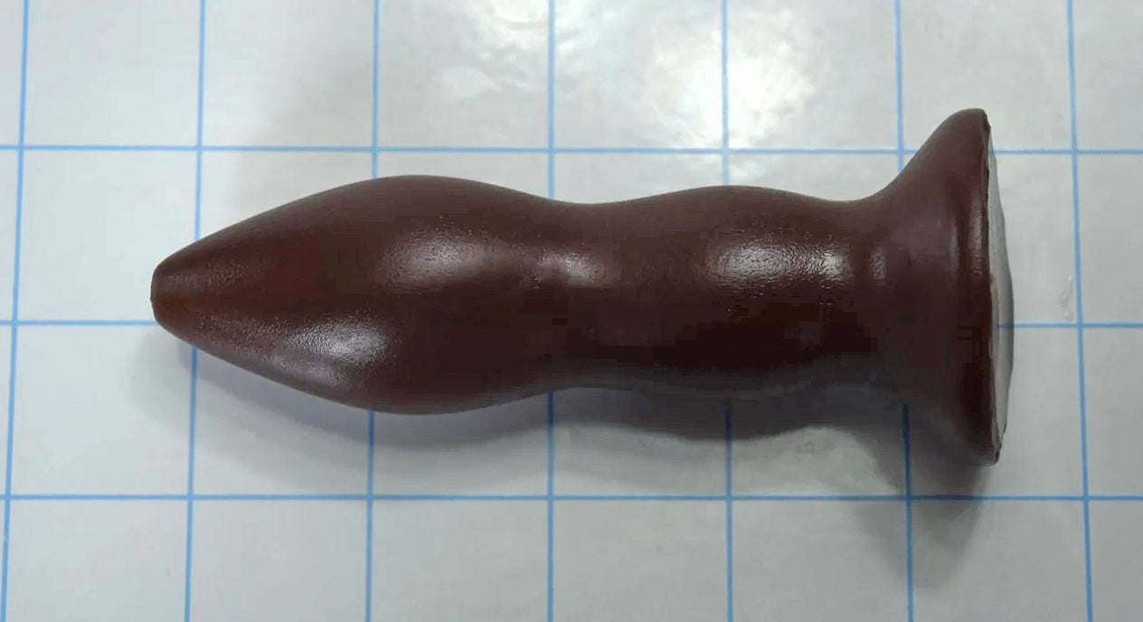 Brown silicone rectal training dilator lying horizontally on a blue grid background.