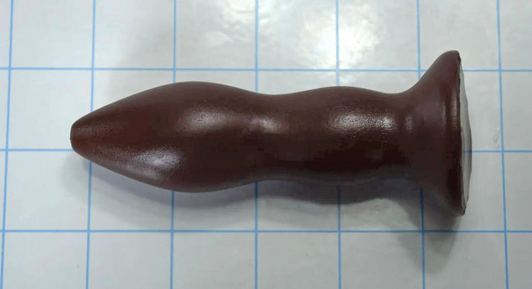 Brown silicone rectal training dilator lying horizontally on a blue grid background.