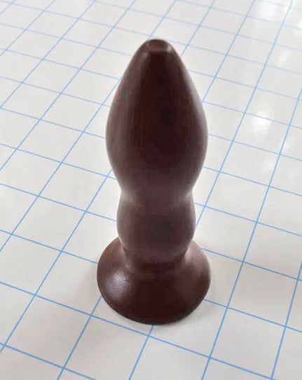 Brown silicone rectal training dilator standing upright on a blue grid background.