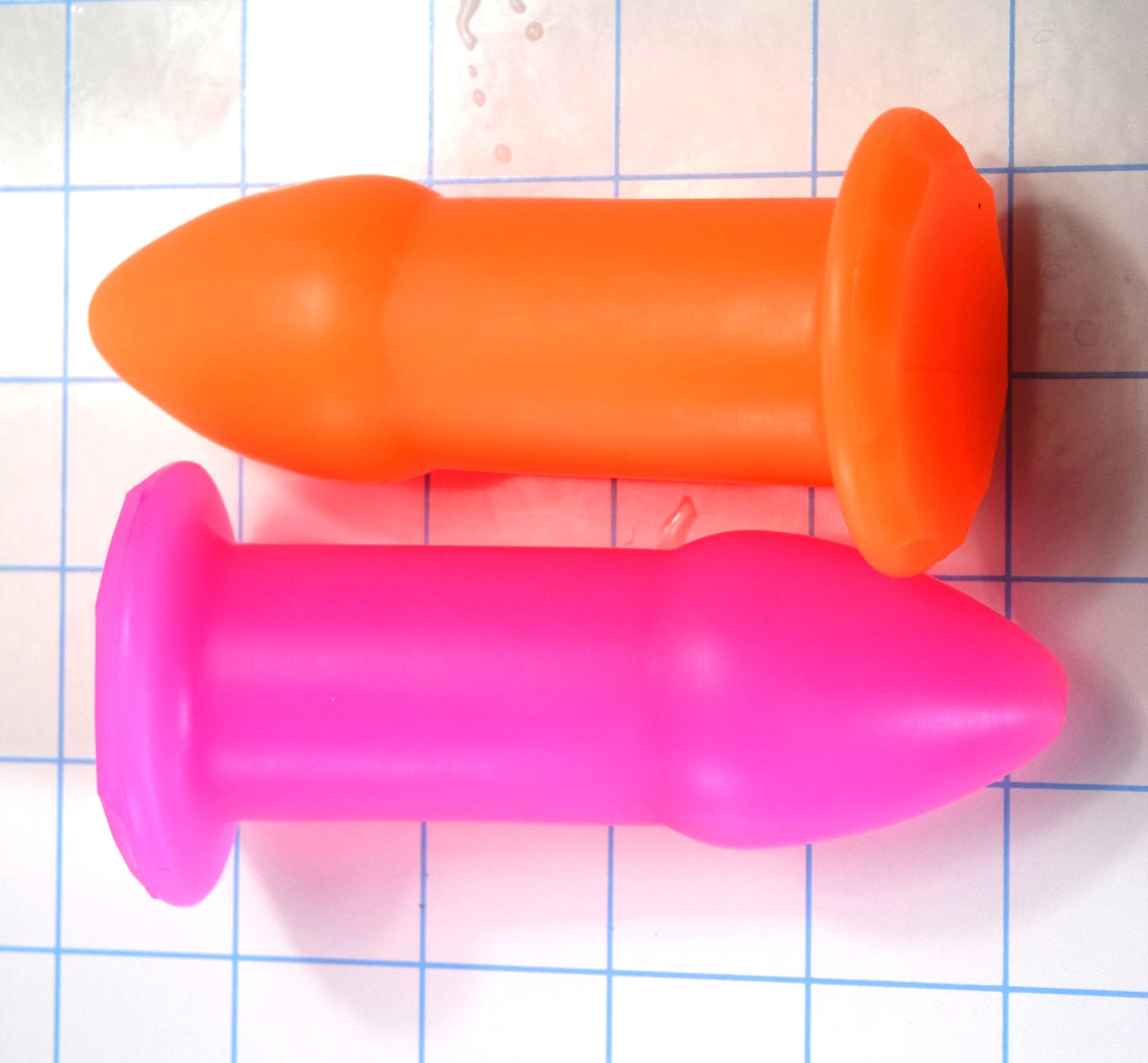 Orange and pink silicone rectal training dilators lying horizontally side-by-side on a blue grid background.