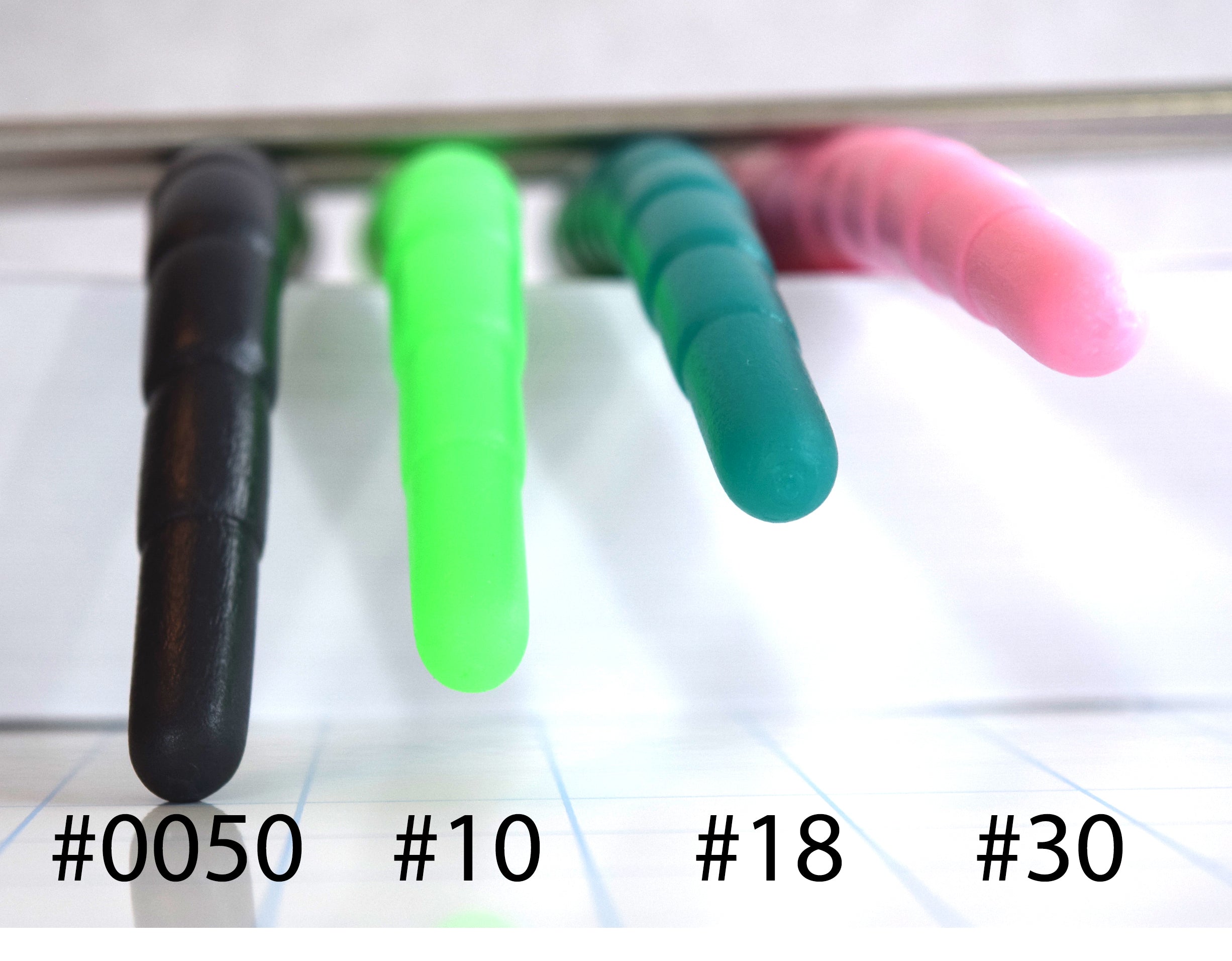 Detailed front view comparison of four silicone dildos in different colors (dark green, light green, teal, pink), labeled #0050, #10, #18, and #30, showing the variation in droop and flexibility.