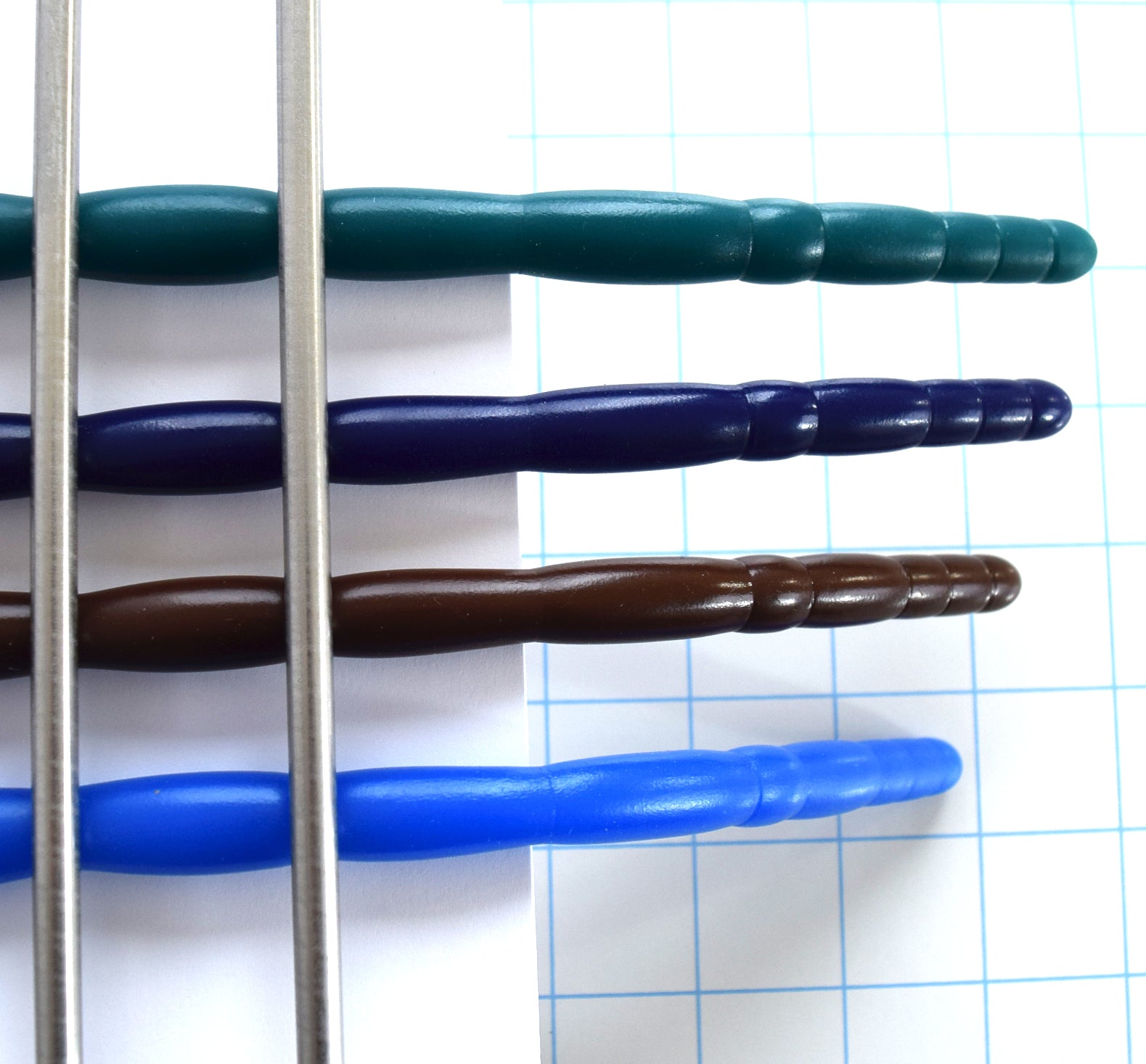 Top-down view of teal, purple, brown, and light blue silicone urethral sounds clamped horizontally to demonstrate firmness and rigidity over grid paper