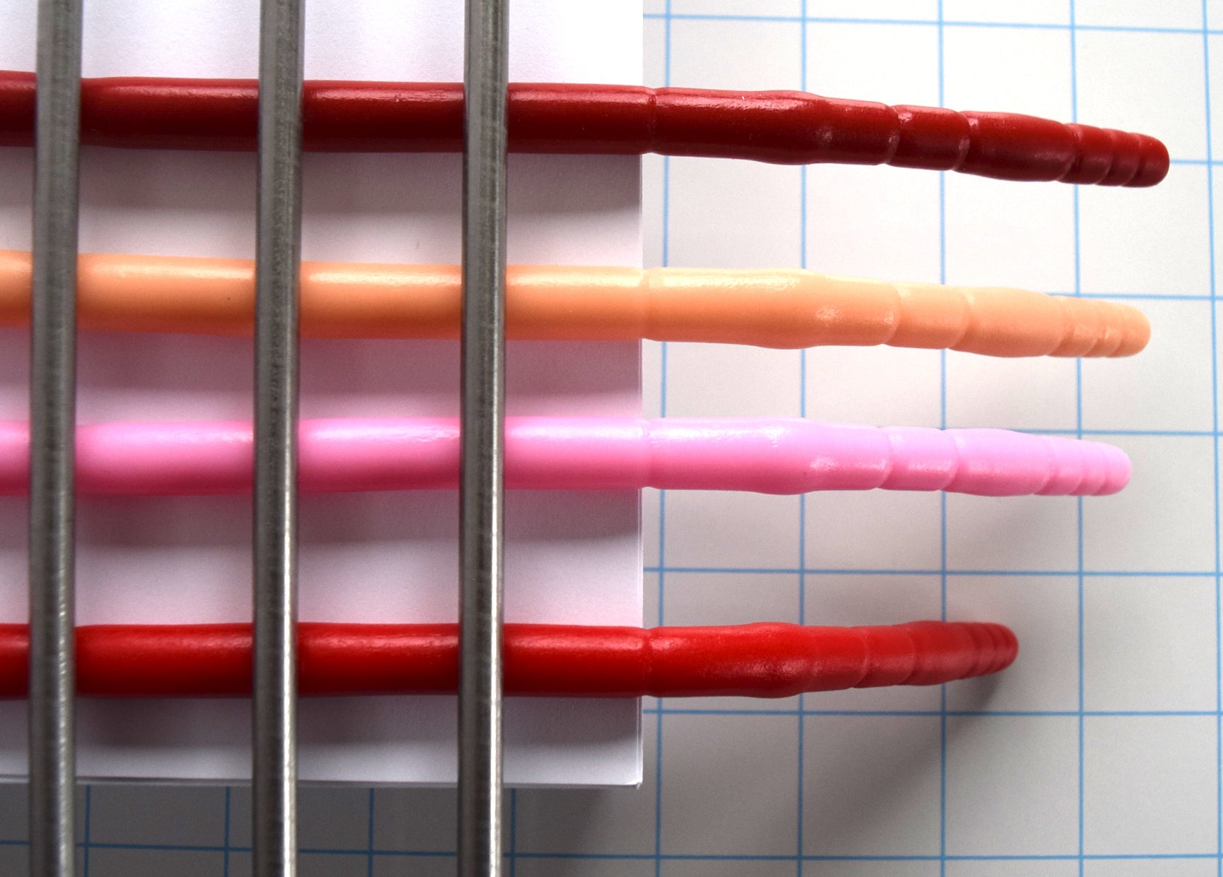 Top-down close-up view of four silicone urethral sounds clamped in place extending over a blue grid paper background to demonstrate firmness and flexibility