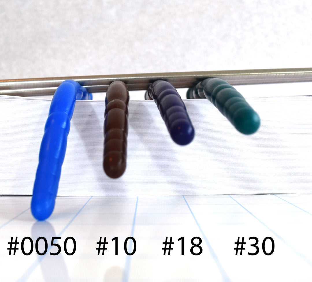 Top-down view comparison of silicone firmness levels #0050, #10, #18, and #30 drooping from a clamp to illustrate flexibility, with numbered labels on a white background.