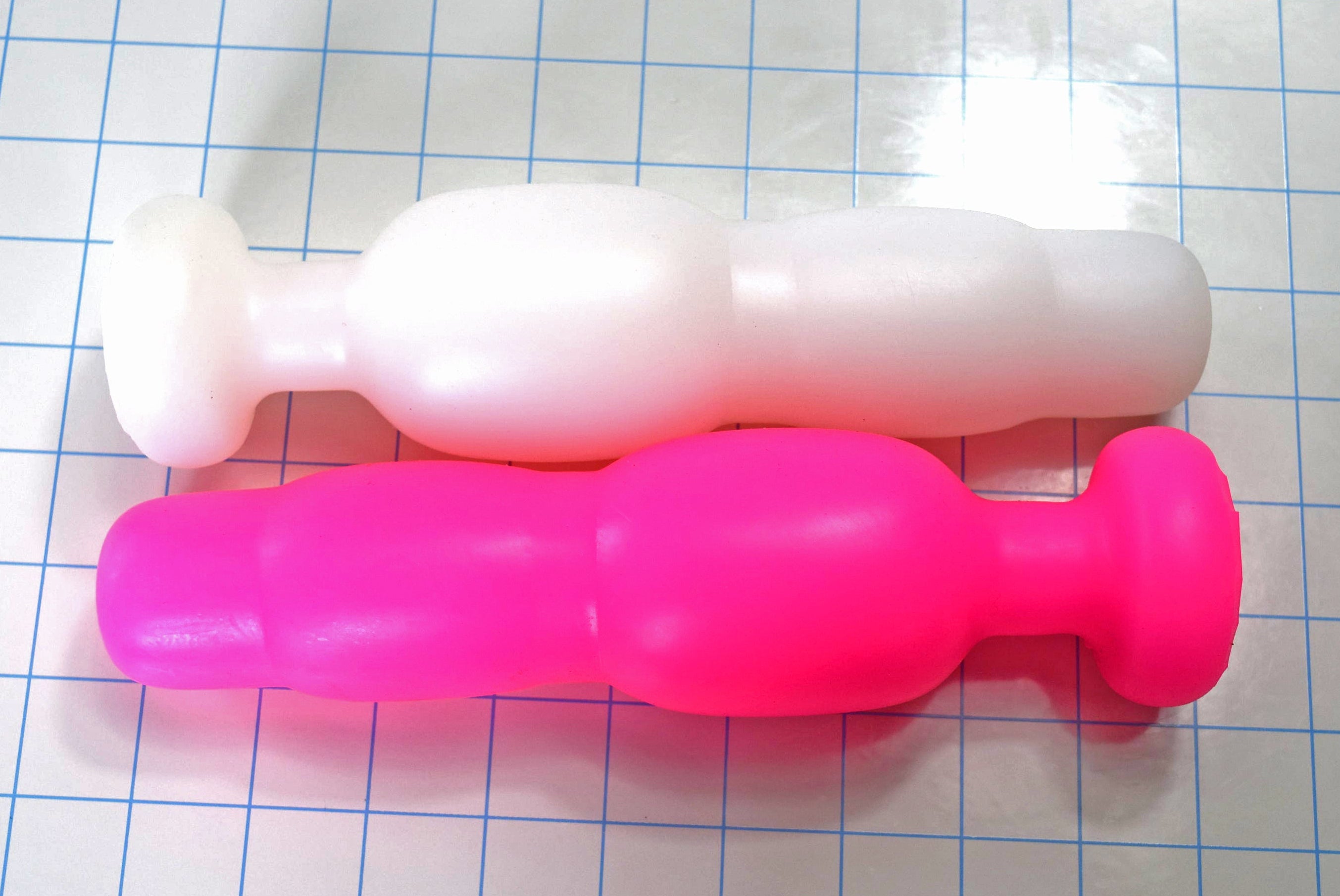 Pink and translucent white stepped silicone retention enema nozzles lying side-by-side on a blue grid background.