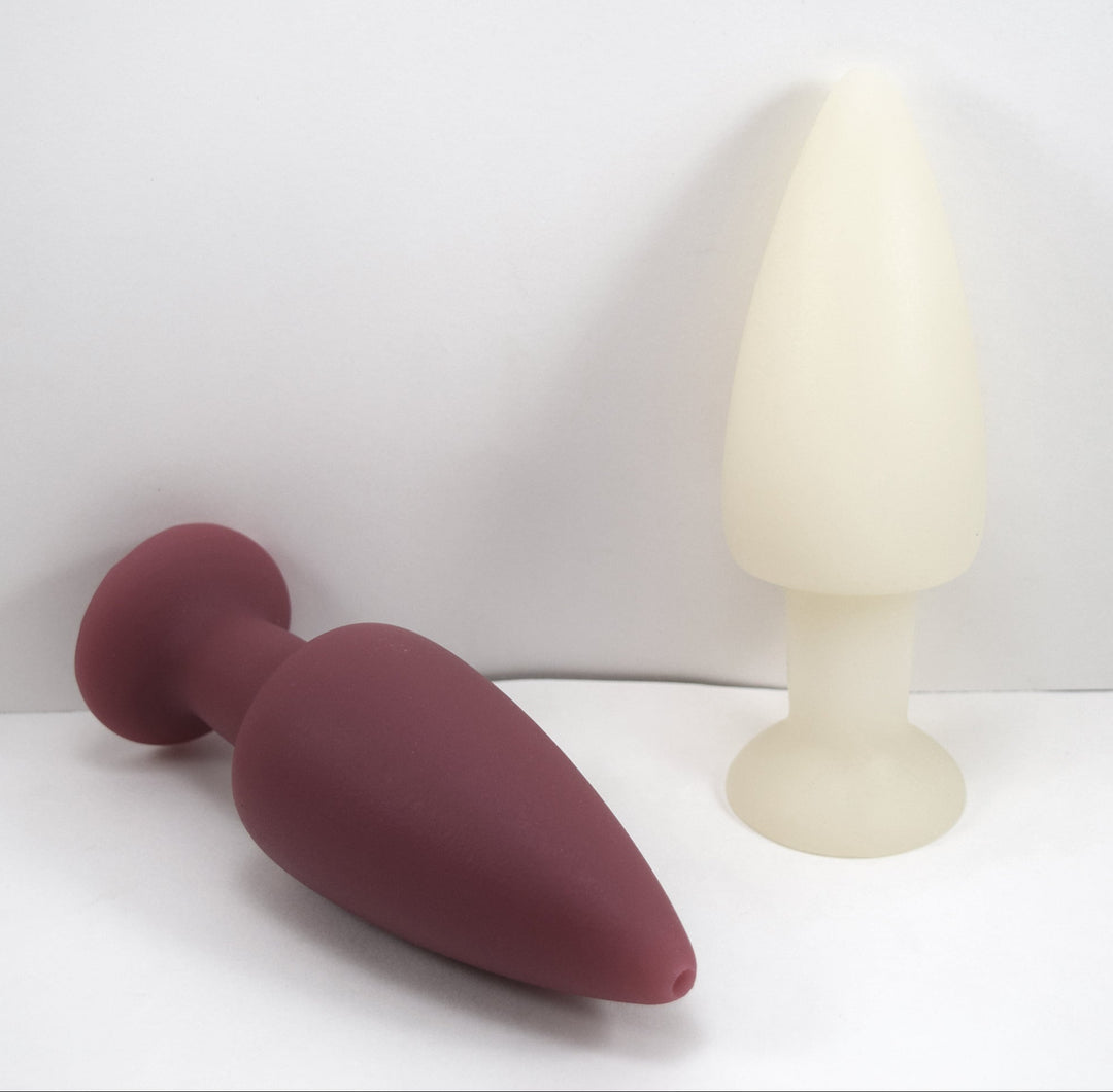 Maroon silicone retention enema nozzle lying down beside an upright white nozzle on a plain white background.