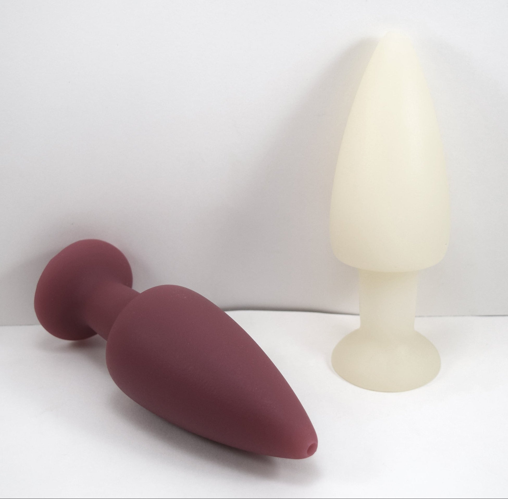 Maroon silicone retention enema nozzle lying down beside an upright white nozzle on a plain white background.